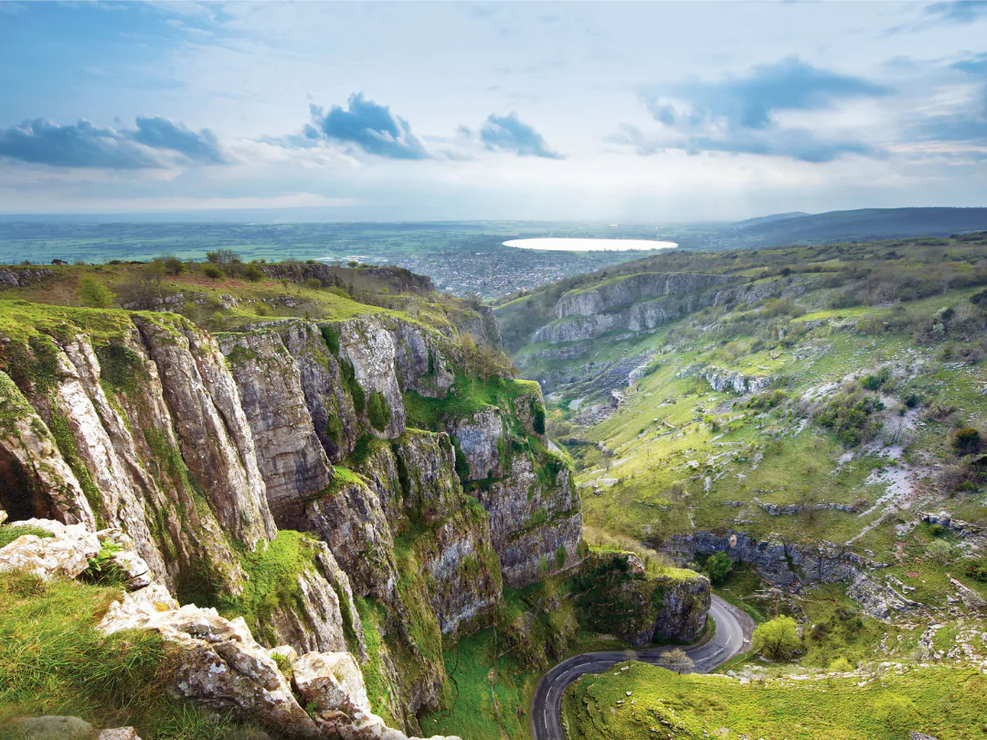 iew of the Mendip Hills, featuring dramatic limestone cliffs, green slopes, and a winding road, with a lake and distant town visible under a partly cloudy sky.