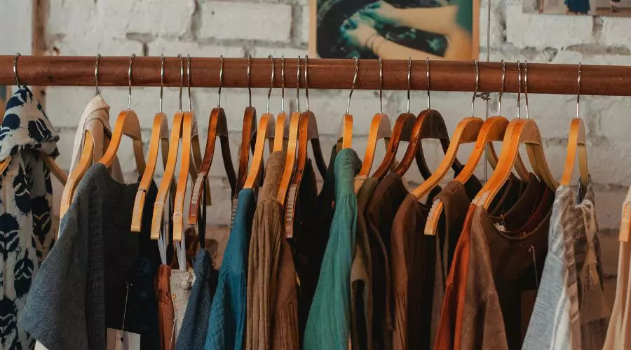A rack displaying various colourful and unique garments on wooden hangers against a rustic brick wall background.