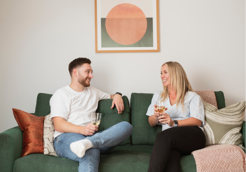 A man and a woman sitting on a green couch, smiling and holding drinks while engaging in conversation. The background features a framed abstract artwork with a circular design.