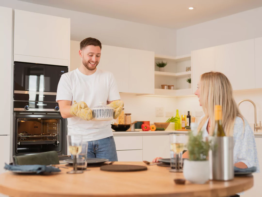 A man smiling in the kitchen holding a hot dish with oven mitts and placing on a kitchen table. Woman is sitting and smiling at the dinner table.