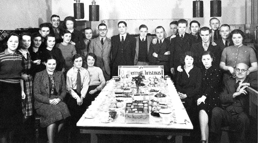 A black and white photograph of a large group of smiling men and women sat around a table of hot drinks and cakes in business attire.