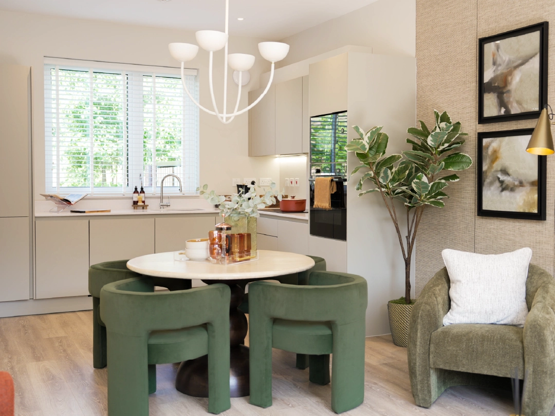 A modern, bright kitchen-dining area at The Coanda Show Home, featuring a round table with green chairs, modern appliances, and large windows with greenery visible outside.