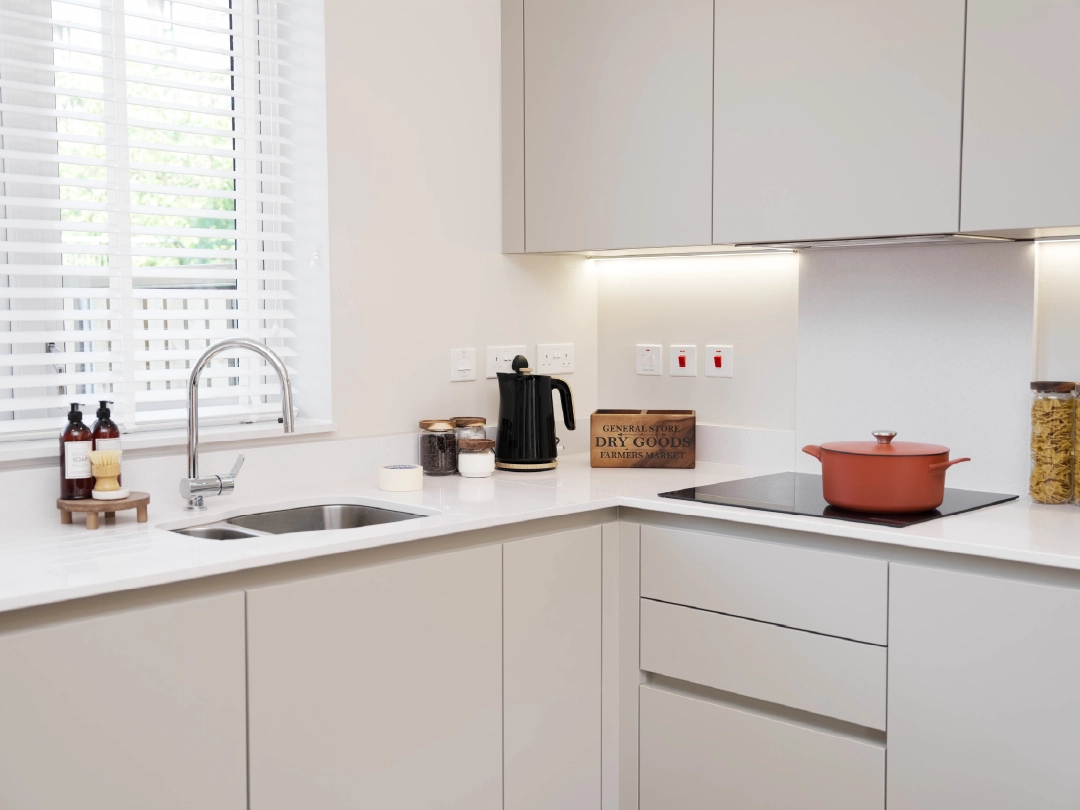 Modern kitchen at Brabazon with white cabinetry, a sink, kettle, and a red pot on the stovetop, featuring natural light and organized shelves.