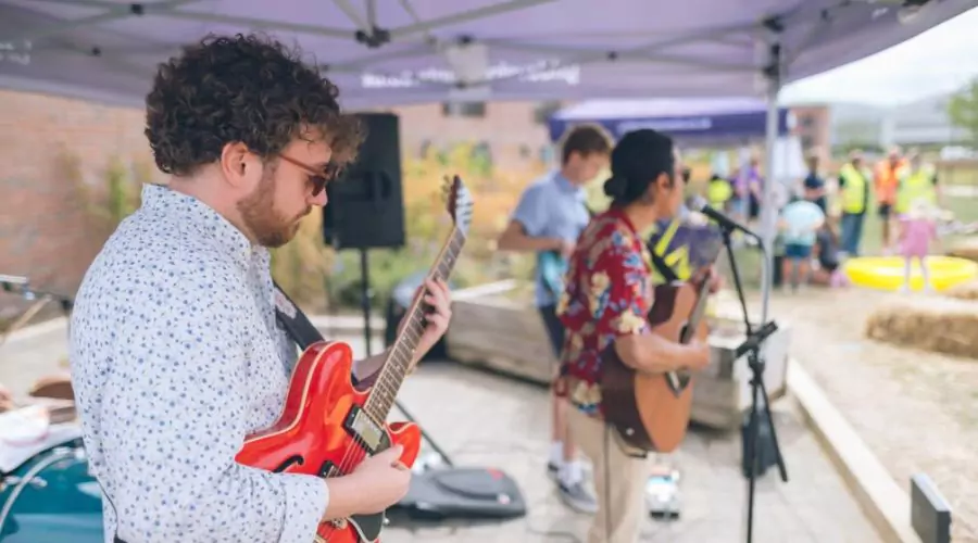 A band performs at an outdoor event at Brabazon, featuring a guitarist with a red guitar and a singer playing an acoustic guitar in a floral shirt.