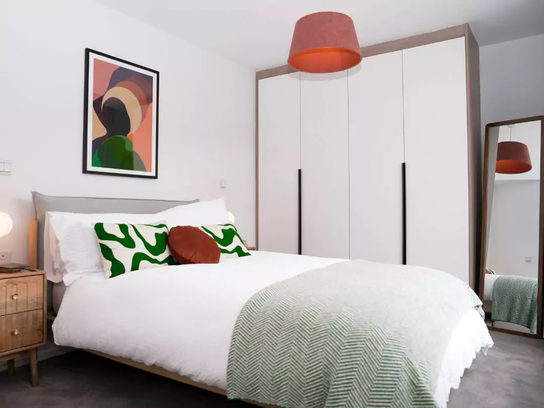 Light and modern primary bedroom, showcasing a double bed with white linen, assorted green pillows, a green throw. Behind is built in white wardrobe and a free standing full length mirror. 