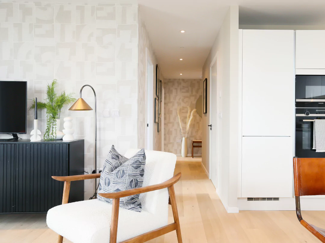 Modern living space with a white armchair, black sideboard, gold floor lamp, and a hallway leading to the kitchen with sleek white cabinetry.