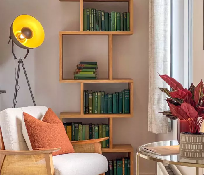 Light reading nook, with a white single arm chair and orange pillow. Behind is a modern shelf full of varying green books. 